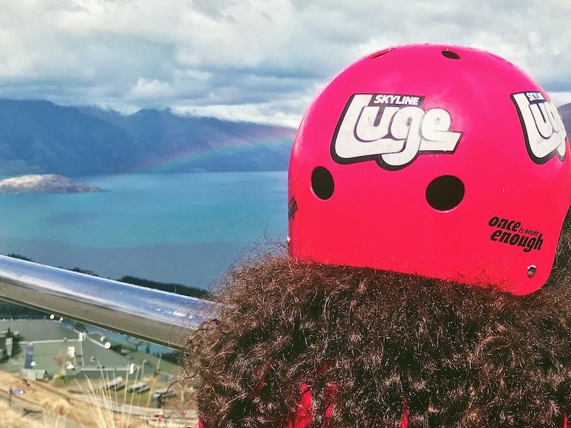 Girl wearing a red Skyline Luge helmet watches a rainbow form over Queenstown.